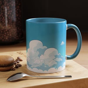 May include: A blue ceramic mug with a cloud design. The mug features a light blue handle and a sky-blue background with white cartoon clouds. A silver spoon and a pastry are on a wooden surface.
