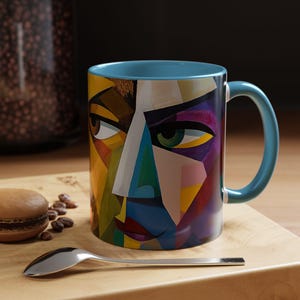 May include: A blue ceramic mug with a colorful abstract design of a face. The face is made up of geometric shapes in shades of yellow, blue, green, purple, and pink.