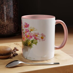 May include: White ceramic mug with a pink interior and handle. The mug features a watercolour painting of pink and white daisies in a grey vase. A silver spoon and a macaron are on a wooden surface.