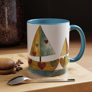 May include: A white ceramic mug with a blue rim and handle features a watercolor illustration of two sailboats with red hearts. The sailboats are yellow, green, and blue.