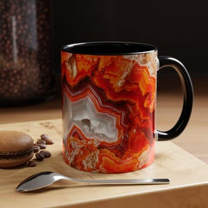 Peut inclure: Une tasse à café noire avec un motif d'agate coloré. Le motif présente des nuances d'orange, de rouge et de blanc.