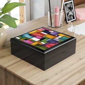 May include: A black wooden box with a colourful geometric design on the lid. The lid features a mosaic of rectangular and square shapes in various colours, including red, blue, green, and yellow. The box is on a wooden surface.