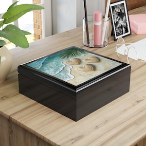 May include: Black wooden box with a hinged lid. The lid features a colorful image of three hearts drawn in the sand on a beach with a palm tree and ocean waves.