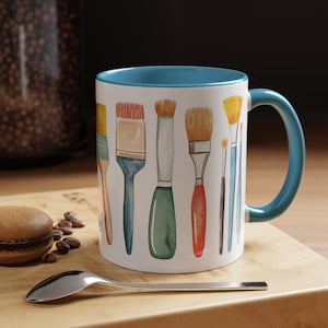 May include: White ceramic mug with a blue interior and handle, decorated with watercolor illustrations of paintbrushes in various sizes and colors. A silver spoon and a pastry are on a wooden surface.