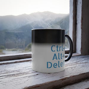 May include: A black and white color-changing mug with the text "Ctrl+Alt+Delete" in blue. The mug is sitting on a wooden surface in front of a window.