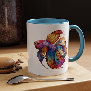 May include: A white ceramic mug with a blue rim and handle features a colorful illustration of a betta fish. The fish has a red, yellow, blue, and purple body with flowing fins.