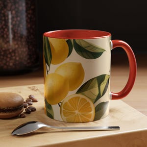 May include: A red and white ceramic mug with a lemon print. The mug has a red rim and handle. The lemon print features yellow lemons and green leaves.