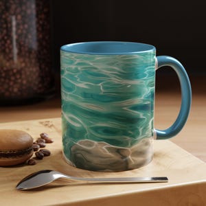 May include: A ceramic coffee mug with a blue handle and interior. The exterior features a turquoise water design with white highlights. The mug sits on a wooden cutting board with a silver spoon, a macaron, and coffee beans.