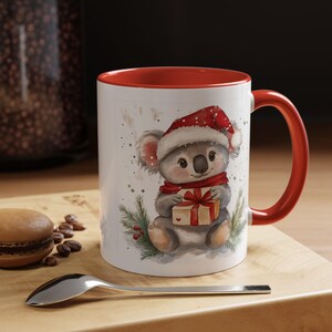 May include: White ceramic mug with a red interior and handle. The mug features a watercolor illustration of a koala wearing a Santa hat and scarf, holding a gift. The design includes sprigs of greenery and a festive theme.