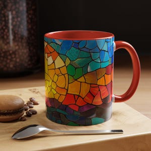 Op de afbeelding: Een rode koffiemok met een kleurrijk mozaïekontwerp. Het mozaïekpatroon heeft tinten blauw, groen, geel, oranje en rood.