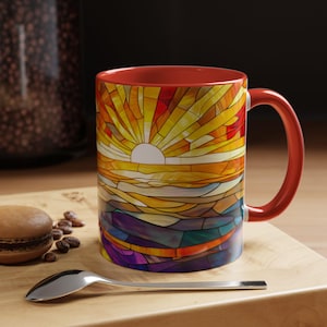 Puede incluir: Una taza de cerámica roja con un diseño de vitral de una salida del sol sobre un paisaje. La taza tiene un interior blanco y una asa.