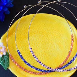 Könnte beinhalten: Drei silberne Draht-Halsketten mit bunten Perlen. Die Halsketten sind übereinander gelegt. Die obere Halskette hat rosa Perlen, die mittlere Halskette hat blaue Perlen und die untere Halskette hat orange Perlen.