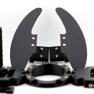 May include: A collection of black automotive parts, including paddle shifters with a carbon fiber weave pattern, a textured cylindrical grip, and various mounting components. The parts are arranged against a white background.