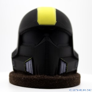 May include: A black helmet with a yellow stripe on top. The helmet has a visor and a textured surface. The helmet is sitting on a brown base.