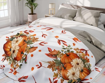 May include: White blanket featuring a fall-themed design with orange pumpkins, white daisies, and autumn leaves. The blanket is decorated with orange leaves and dots, creating a cozy, seasonal aesthetic. The blanket is on a bed with neutral-colored bedding.