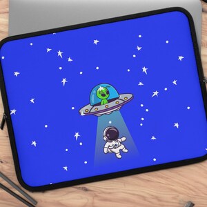 May include: A blue laptop sleeve featuring a cartoon illustration of a green alien in a UFO beaming down on an astronaut. The background is a starry night sky with white stars and dots. The sleeve has a black border.