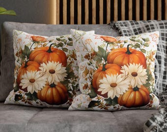Puede incluir: Dos cojines decorativos con temática otoñal. Cada cojín presenta un diseño de calabazas naranjas y margaritas blancas con hojas verdes sobre un fondo blanco. Los cojines están sobre un sofá gris.