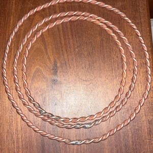 Op de afbeelding: Drie cirkelvormige koperdraad kettingen met een gedraaid touwontwerp. Elke ketting is gevormd uit een doorlopende streng van metalen koperdraad met een duidelijke gevlochten textuur. Een kleine bijpassende koperen armband ligt erboven.