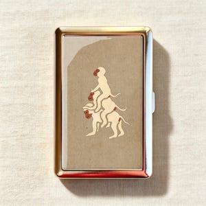 Puede incluir: Una pitillera rectangular de metal con acabado dorado pulido. La parte frontal presenta una inserción de color tostado con una ilustración gráfica de tres monos de color crema apilados verticalmente uno sobre otro con caras de color marrón rojizo.