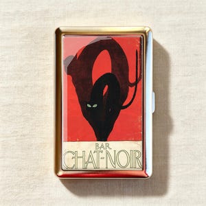 May include: A rectangular metal case with a gold-toned frame featuring a graphic print of a black cat with green eyes against a vibrant red background. The lower portion displays the text BAR CHAT-NOIR in black lettering on a cream-colored base.