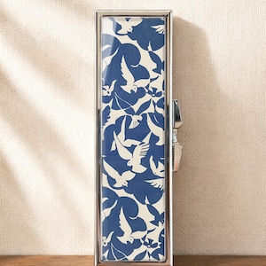 May include: A rectangular metal lipstick case with a polished silver frame and a hinged side closure. The front panel features a repeating pattern of white birds flying against a solid dark blue background, creating a high-contrast graphic design.