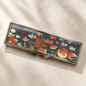 Peut inclure: Un étui rectangulaire en métal avec un couvercle à charnière présentant un fond bleu foncé orné d'illustrations de poissons rouges orange, rouges et noirs, de plantes aquatiques vertes et d'un emblème ovale rouge central avec du texte japonais.