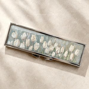 Peut inclure: Un étui rectangulaire en métal argenté doté d'un panneau incrusté représentant une peinture à l'huile impressionniste de tulipes blanches sur un fond vert sauge atténué. L'étui possède un cadre métallique poli et un mécanisme de fermeture.