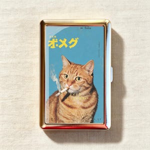 May include: A rectangular gold-toned metal cigarette case featuring a graphic print of an orange tabby cat with a lit cigarette in its mouth. The background is light blue with yellow Japanese text at the top and smaller white text below the cat.