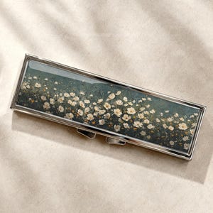 Op de afbeelding: Een rechthoekige metalen etui met een gepolijste zilveren afwerking, voorzien van een print van witte wilde bloemen verspreid over een donkere, gedempte groene achtergrond. De etui heeft een scharnierend deksel en een slanke, langwerpige vorm.
