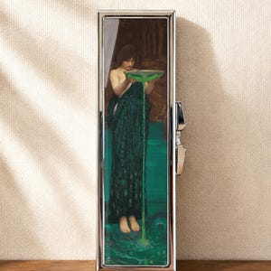Op de afbeelding: Een rechthoekige zilverkleurige metalen behuizing met een reproductie van het schilderij Circe Invidiosa van John William Waterhouse. Het kunstwerk toont een figuur in een donkergroene jurk die een felgroene vloeistof uit een kom giet.