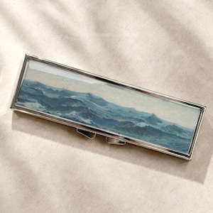 Op de afbeelding: Een rechthoekige metalen doos met een gepolijste zilveren afwerking, voorzien van een ingelegde schildering van een woelige oceaan met donkerblauwe en witte golven onder een bleke lucht. De doos heeft een scharnierend deksel met een sluiting.