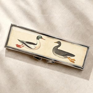 Op de afbeelding: Een rechthoekige zilverkleurige metalen doos met een illustratie van twee eenden op een crèmekleurige achtergrond. De linker eend heeft een zwarte kop, een wit lichaam en rode poten, terwijl de rechter eend donkerbruin is met oranje poten.