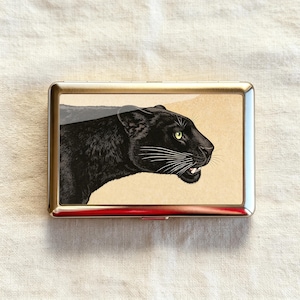 Peut inclure: Un étui métallique rectangulaire avec un cadre argenté poli présentant une illustration détaillée d'une panthère noire de profil. La panthère a des yeux jaunes, des moustaches blanches et des dents visibles sur un fond beige clair.