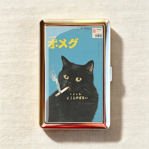 Op de afbeelding: Een rechthoekig sigarettendoosje van goudkleurig metaal met een grafische print van een zwarte kat met gele ogen die een brandende sigaret in de bek houdt. De blauwe achtergrond bevat gele Japanse tekst en een kleine barcode rechtsboven.