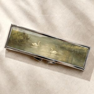 Peut inclure: Un étui rectangulaire en métal argenté doté d'un panneau incrusté représentant deux cygnes blancs nageant sur un étang aux tons verts et bruns, entourés de hautes herbes sous un ciel doux et nuageux.
