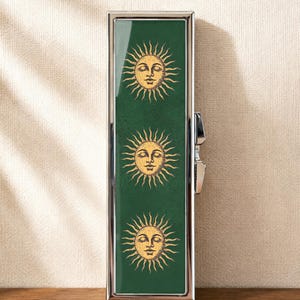 Peut inclure: Un étui rectangulaire en métal argenté avec un panneau vert foncé sur le devant. Le panneau est orné de trois illustrations identiques de soleils dorés, représentant chacun un visage aux yeux fermés avec des rayons rayonnants.