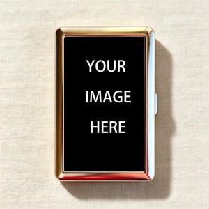 May include: A rectangular metal cigarette case with a polished gold-tone finish and a central black panel. The black panel features the white text YOUR IMAGE HERE, indicating a customizable surface for personal designs or photographs.