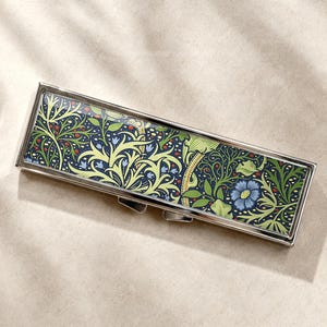 Peut inclure: Un pilulier rectangulaire en métal doté d'une incrustation brillante ornée d'un motif botanique dense de fleurs bleues, de feuillage vert et de petites baies rouges sur fond bleu marine, le tout dans un cadre en métal argenté poli.