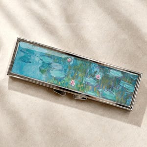 Op de afbeelding: Een rechthoekige metalen houder met een scharnierend deksel voorzien van een gedrukte reproductie van een schilderij van een waterlelievijver. Het kunstwerk toont tinten blauw, groen en taling met kleine roze bloemaccenten en een zilverkleurig frame.