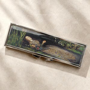 Peut inclure: Un boîtier rectangulaire en métal argenté présentant une reproduction de la peinture Ophélie de John Everett Millais sur le couvercle. L'œuvre représente une silhouette allongée dans un ruisseau entourée de roseaux verts et de fleurs.