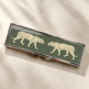 May include: A rectangular metal pill case featuring a dark olive green top panel decorated with two cream-colored tiger illustrations facing each other. The case has a polished silver-tone frame and a hinged lid with a secure clasp mechanism.