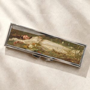 Pode incluir: Um estojo retangular de metal prateado com uma reprodução da pintura Ofélia de John Everett Millais na tampa. A obra retrata uma pessoa com uma túnica branca flutuando num lago rodeado por folhagem verde e flores brancas.