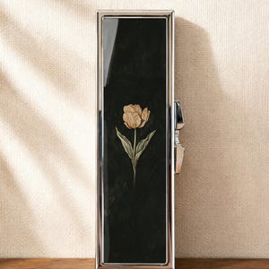 Peut inclure: Un étui rectangulaire en métal argenté doté d'une incrustation noire brillante sur le panneau avant. Au centre de la surface noire se trouve une illustration détaillée d'une tulipe claire unique avec des feuilles vertes. Fermoir latéral.