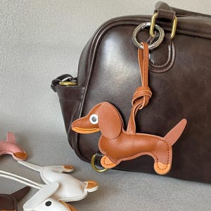 Può includere: Un portachiavi a forma di cane bassotto in pelle marrone con un anello dorato attaccato a un cinturino in pelle marrone. Il cane ha dettagli beige sulle zampe e sulle orecchie. Il portachiavi è appeso a una borsa in pelle marrone.