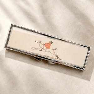 Peut inclure: Un étui rectangulaire en métal argenté présentant une illustration centrale d'un cavalier en veste rouge sur un cheval. L'étui possède un cadre métallique poli et un panneau central lisse de couleur claire illustrant la scène équestre.