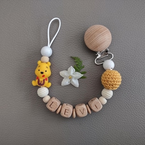 Könnte beinhalten: Ein Schnullerhalter mit einer gelben Bärenfigur, einer weißen Schlaufe und Holzperlen, die den Namen LEVI bilden. Das Design umfasst einen runden Holzclip, eine senfgelbe gehäkelte Perle sowie kleinere weiße und naturfarbene Holzperlen.