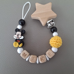 Könnte beinhalten: Ein Schnullerhalter mit einem sternförmigen Holzclip, einer Mickey-Maus-Figur, einer gelben gehäkelten Kugel und Holzbuchstabenperlen, die den Namen MIKE bilden, ergänzt durch kleine weiße und schwarze Perlen an einer weißen Kordelschlaufe.