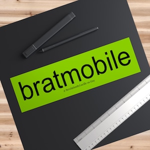 May include: A neon green sticker with the word "bratmobile" in black, along with the Etsy shop name. A black stylus and ruler are also present on a black surface.