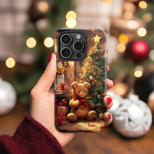 Puede incluir: Una funda para teléfono con una escena navideña que presenta un oso de peluche, un árbol de Navidad decorado y regalos. La funda tiene un nombre personalizado "Helen" en la parte inferior.