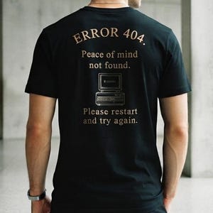 May include: A black short-sleeved t-shirt featuring gold text on the back that reads ERROR 404. Peace of mind not found. Please restart and try again. Below the text is a graphic illustration of a vintage computer monitor and keyboard.